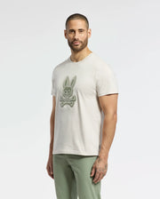 A person with short dark hair and a beard sports the Psycho Bunny MENS SYDNEY GRAPHIC TEE (B6U508F200), a cream Pima cotton t-shirt featuring a bunny skull and crossed bones graphic, paired with light green pants against a plain white background.