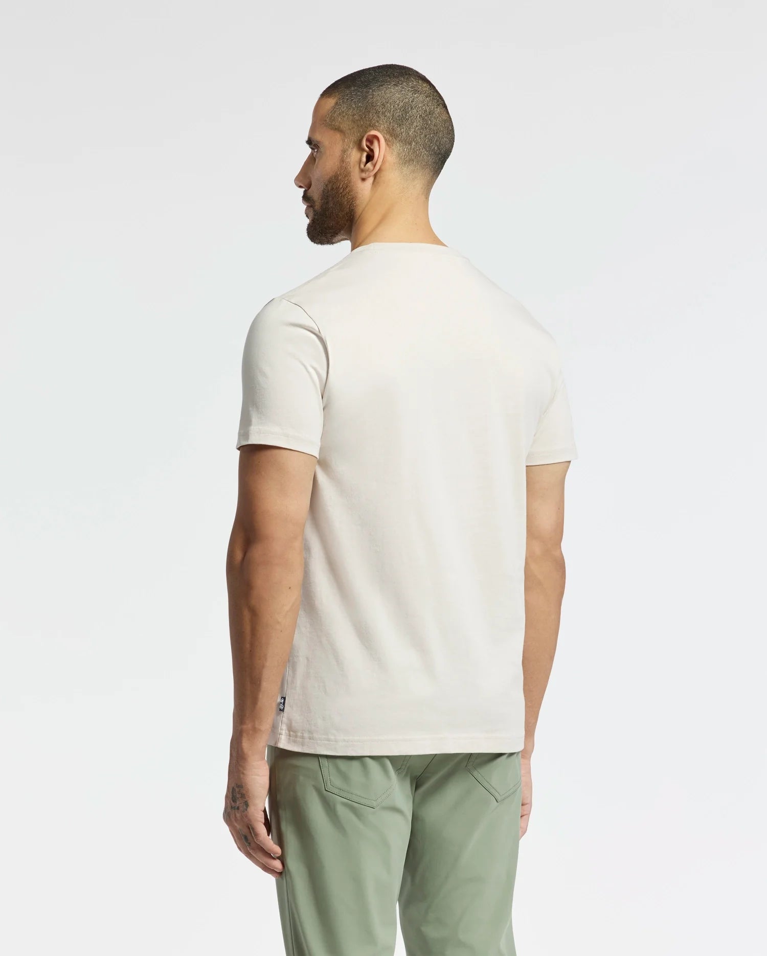 A man, sporting short hair and a beard, stands with his back to the camera. He wears a Psycho Bunny MEN'S SYDNEY GRAPHIC TEE - B6U508F200 made from soft Pima cotton paired with green pants against a neutral backdrop that highlights his understated style.