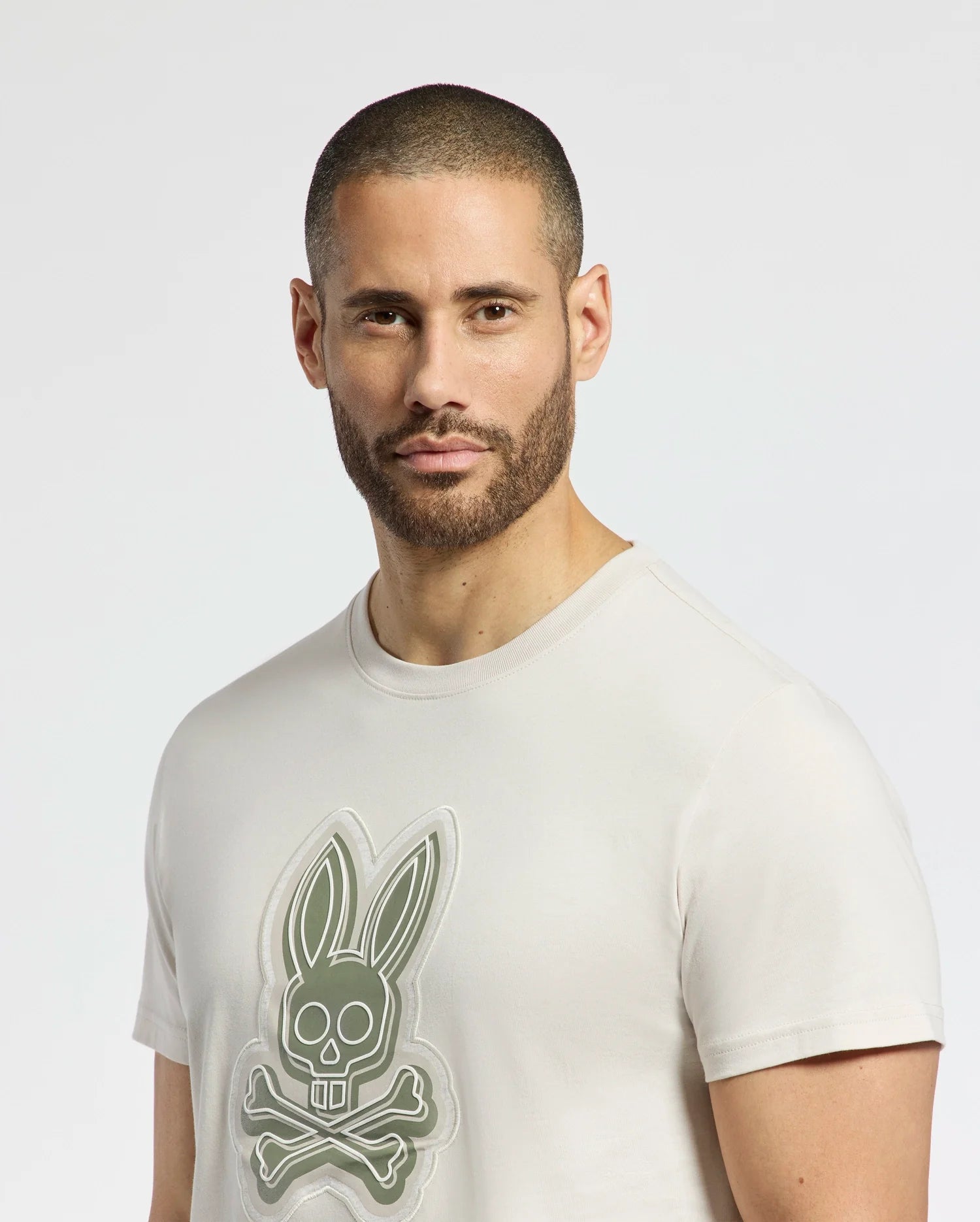 A man with short dark hair and a beard wears the Psycho Bunny's MENS SYDNEY GRAPHIC TEE - B6U508F200, a regular-fit, light-colored T-shirt crafted from soft Pima cotton. It features a bold bunny skull and crossbones graphic on a clean, plain white background.