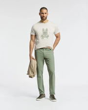 A man wearing the Psycho Bunny MENS SYDNEY GRAPHIC TEE - B6U508F200, a light Pima cotton T-shirt with a bunny graphic, and green pants stands against a plain background. He holds a light jacket and wears matching green sneakers.