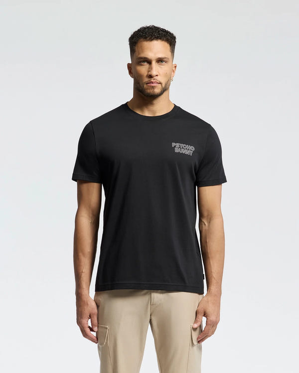 A man in a regular fit Psycho Bunny MENS SYDNEY BACK GRAPHIC TEE, crafted from soft Pima cotton with a small chest print, stands against a plain background. His short curly hair and beard complement his casual look, completed with beige pants.
