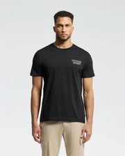 A man in a regular fit Psycho Bunny MENS SYDNEY BACK GRAPHIC TEE, crafted from soft Pima cotton with a small chest print, stands against a plain background. His short curly hair and beard complement his casual look, completed with beige pants.
