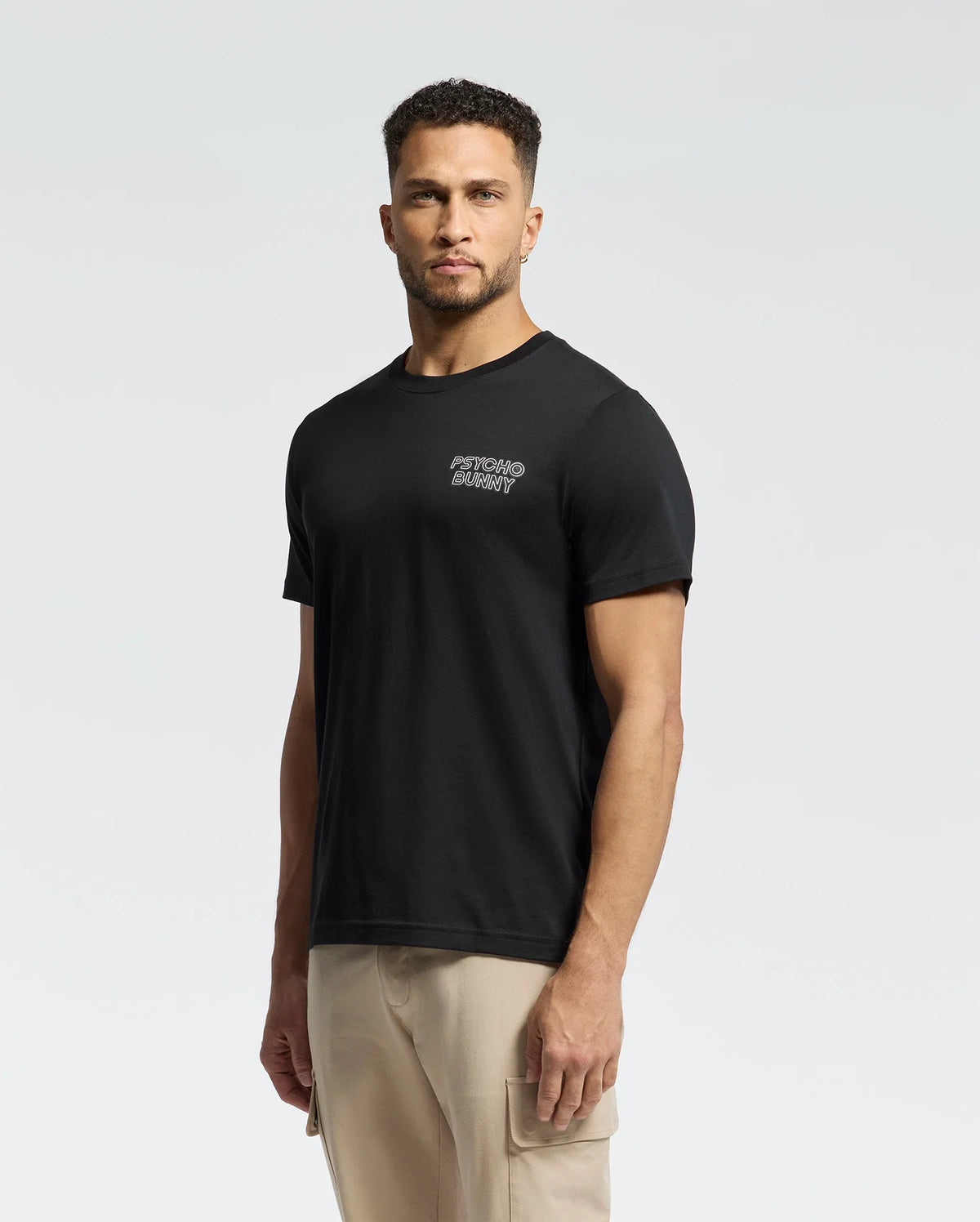 A person in Psycho Bunny's MENS SYDNEY BACK GRAPHIC TEE (B6U509F200), a regular fit, black Pima cotton t-shirt with white water-based print on the upper left, and beige cargo pants stands against a light gray background.