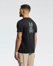 A man with short curly hair, back to the camera, wears a Psycho Bunny MENS SYDNEY BACK GRAPHIC TEE - B6U509F200 in regular fit black Pima cotton with a water-based bunny skull design. He pairs it with beige cargo pants against a plain, light-colored background.