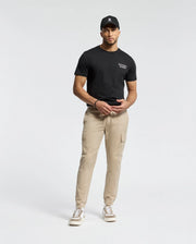 A man in a Psycho Bunny black tee (MENS SYDNEY BACK GRAPHIC TEE - B6U509F200), beige cargo pants, and sneakers stands against a white background. He's wearing a black cap and casually posing with his hands together.