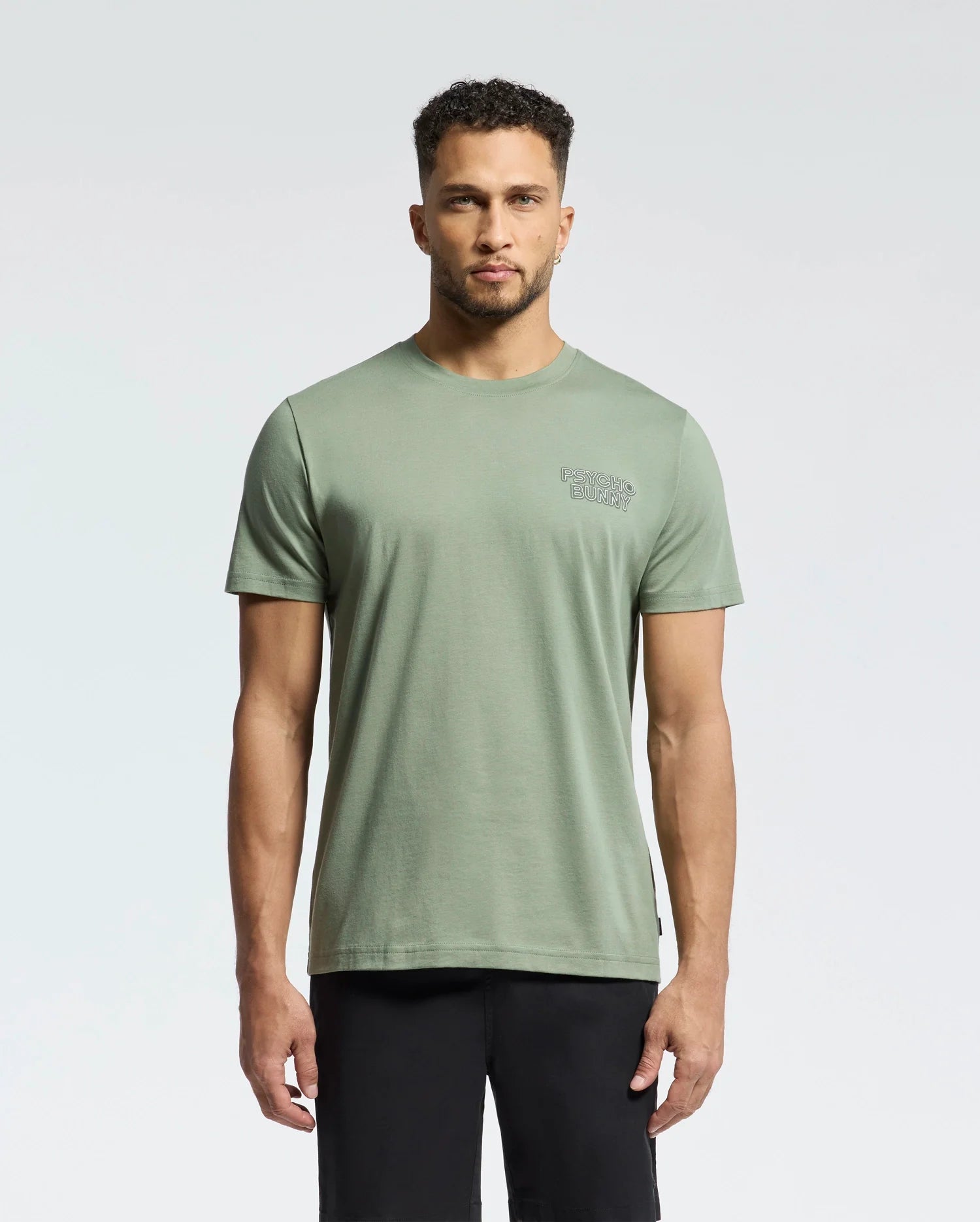 A man in a light green Psycho Bunny MENS SYDNEY BACK GRAPHIC TEE - B6U509F200 and black pants stands against a plain white background. The tee features a small logo on the left chest.