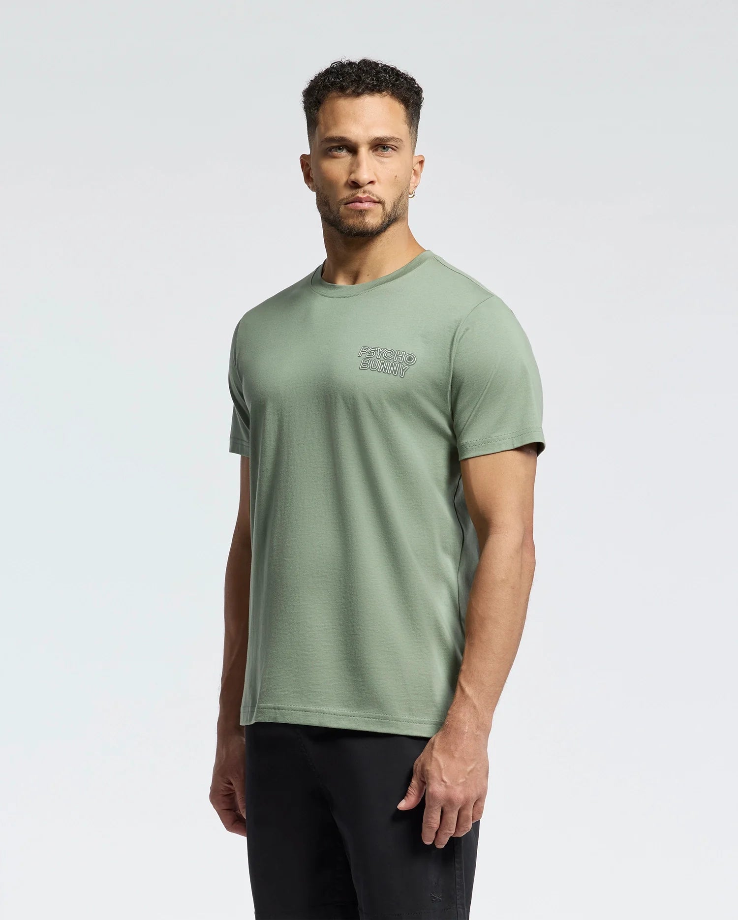 A person in a green Psycho Bunny MENS SYDNEY BACK GRAPHIC TEE - B6U509F200, crafted from soft Pima cotton, paired with black pants, stands against a plain background and gazes at the camera.