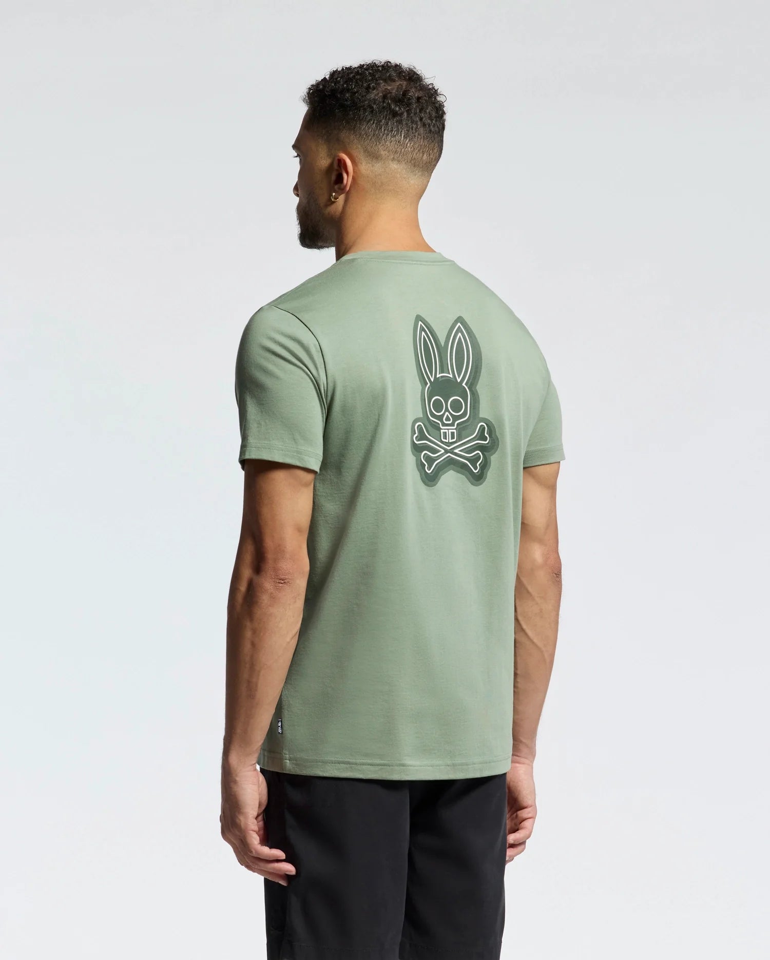 The man, with short curly hair and black pants, is turned away, wearing the Psycho Bunny Men's Sydney Back Graphic Tee - B6U509F200. This light green Pima cotton T-shirt features a skull and crossbones graphic with bunny ears on the back against a plain light gray backdrop and has a classic crewneck style.