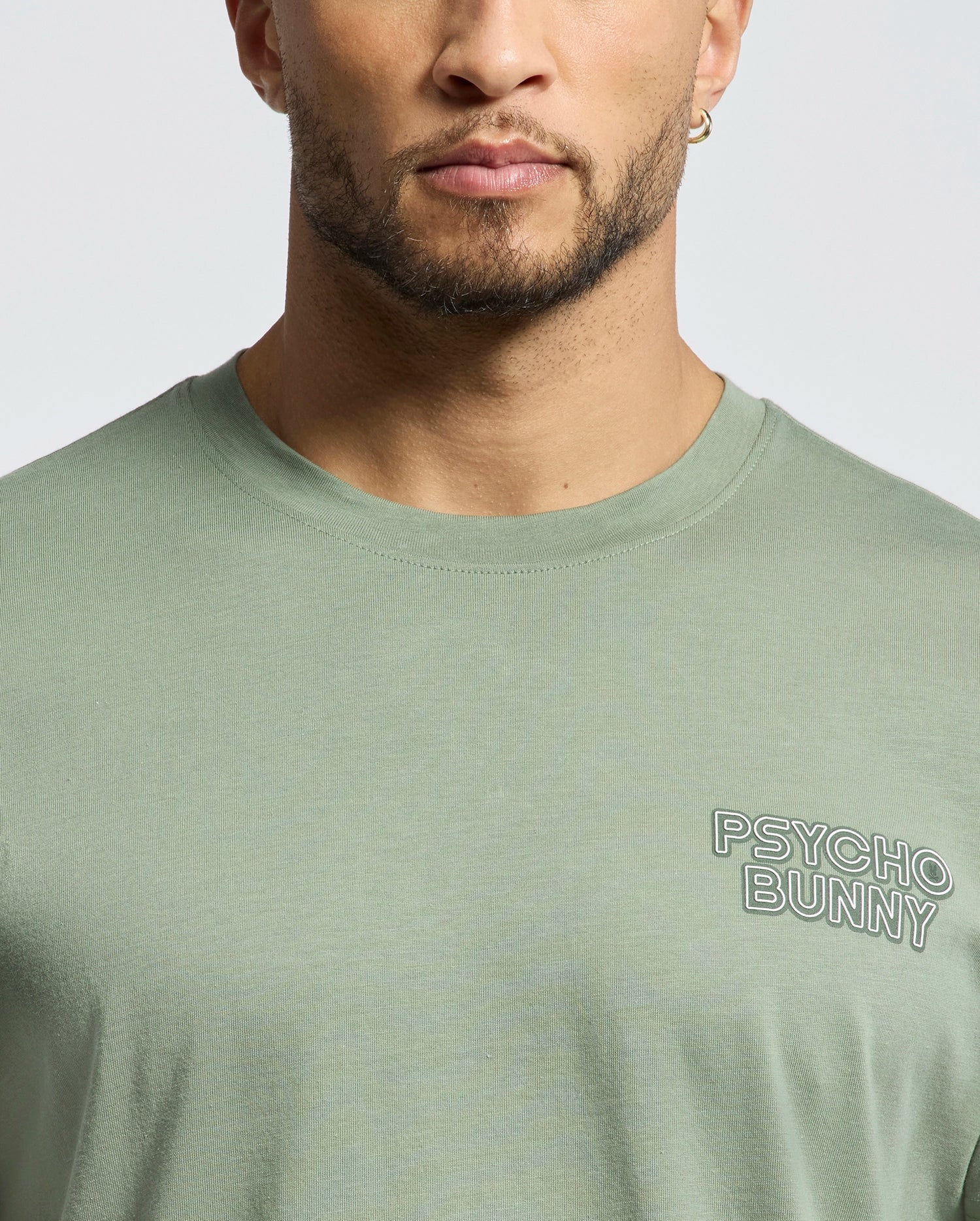 A person sports the "Psycho Bunny" MENS SYDNEY BACK GRAPHIC TEE - B6U509F200 in light green, showcasing the logo on a Pima cotton crewneck. The image captures their face, neck, and upper shirt, highlighting a blend of casual style and comfort.