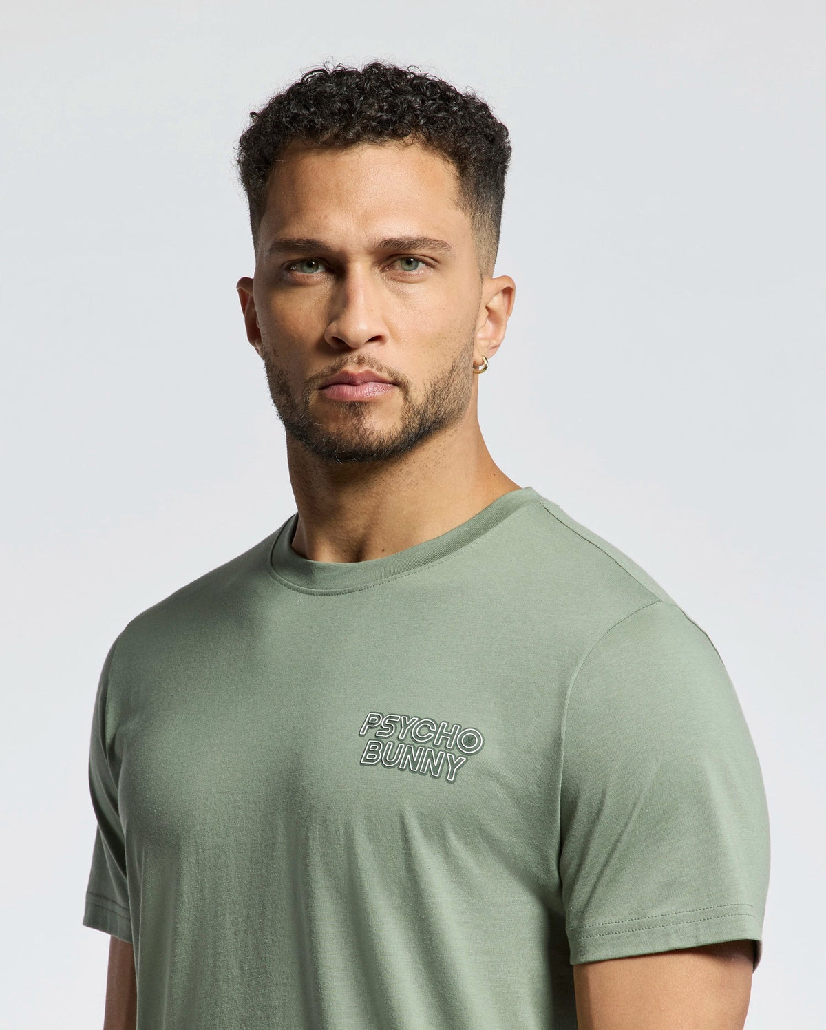 A man in a light green "MENS SYDNEY BACK GRAPHIC TEE - B6U509F200" by Psycho Bunny, crafted from soft Pima cotton, with short curly hair and an earring, looks directly at the camera against a plain backdrop.