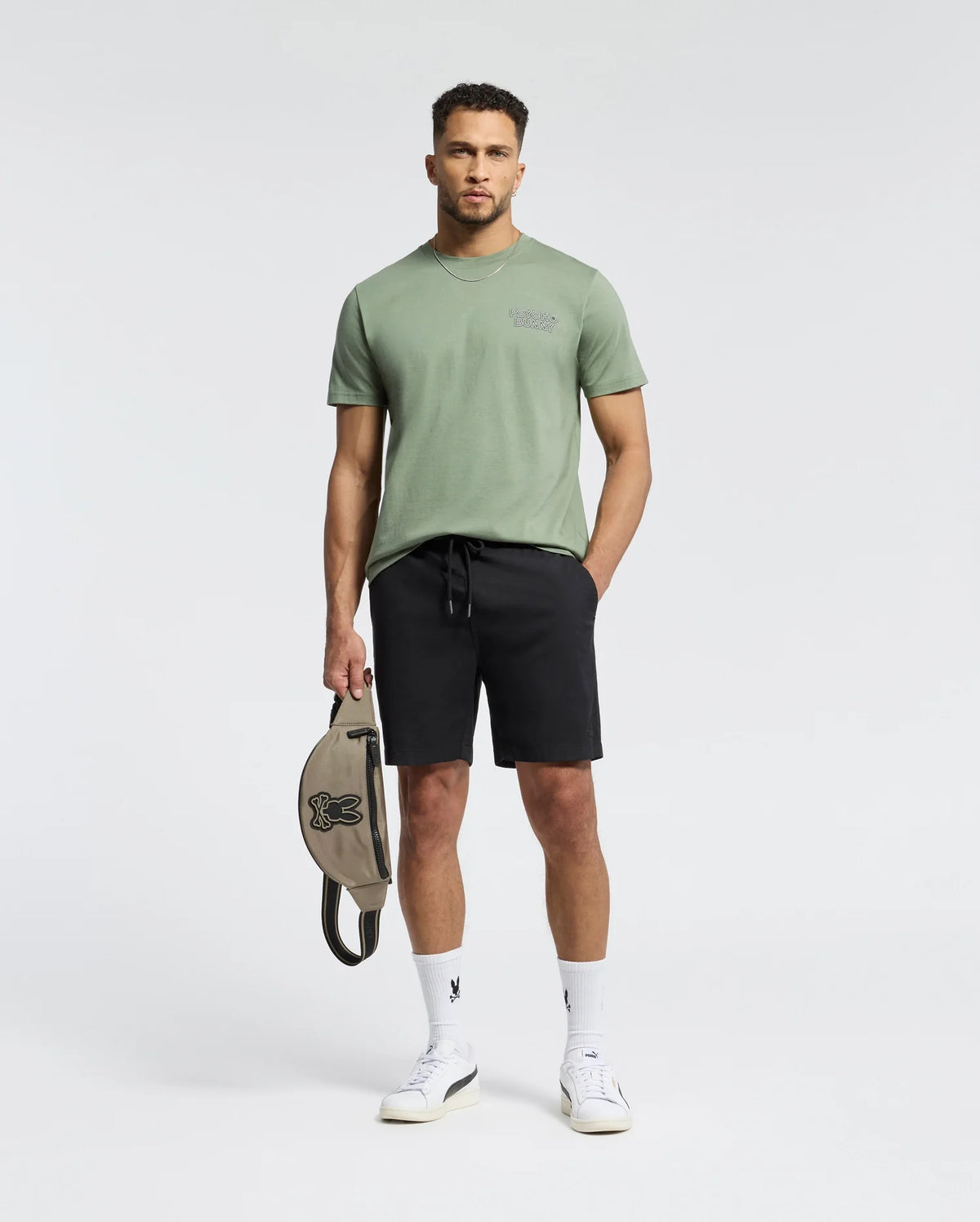 A man in a Psycho Bunny "MENS SYDNEY BACK GRAPHIC TEE - B6U509F200" stands wearing a Pima cotton green T-shirt with a subtle back graphic, black shorts, and holds a bag with a bunny logo. He completes his look with white sneakers and socks against a simple white background.
