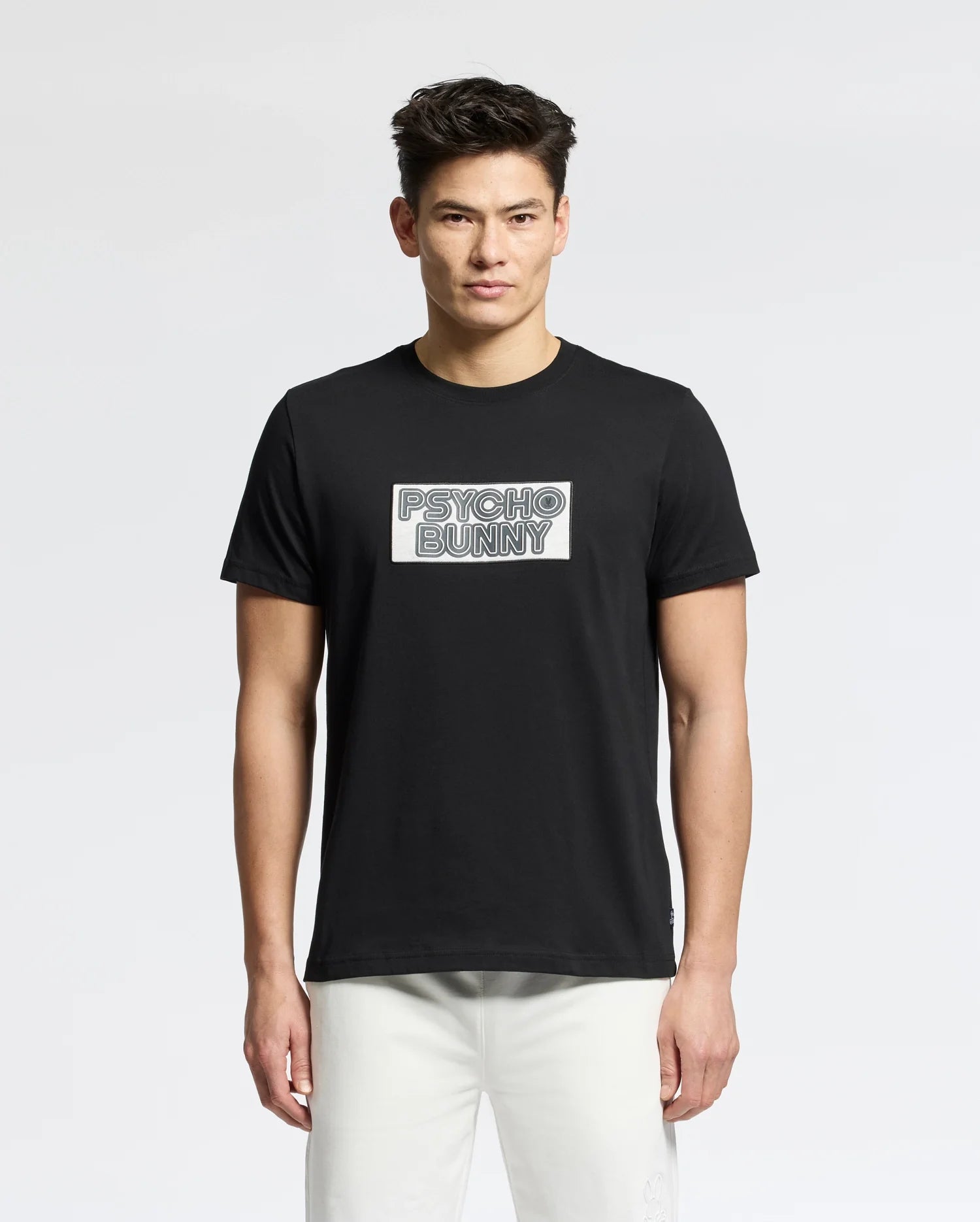 A person stands against a white background wearing light-colored pants and a black Psycho Bunny MENS SYDNEY WORDMARK GRAPHIC TEE - B6U510F200, featuring a water-based print logo. They have short dark hair and look straight ahead, showcasing their modern style.