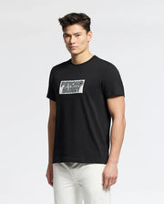 A person in a Psycho Bunny MEN'S SYDNEY WORDMARK GRAPHIC TEE (Black, Pima cotton, water-based print) and white pants stands against a pristine white background.