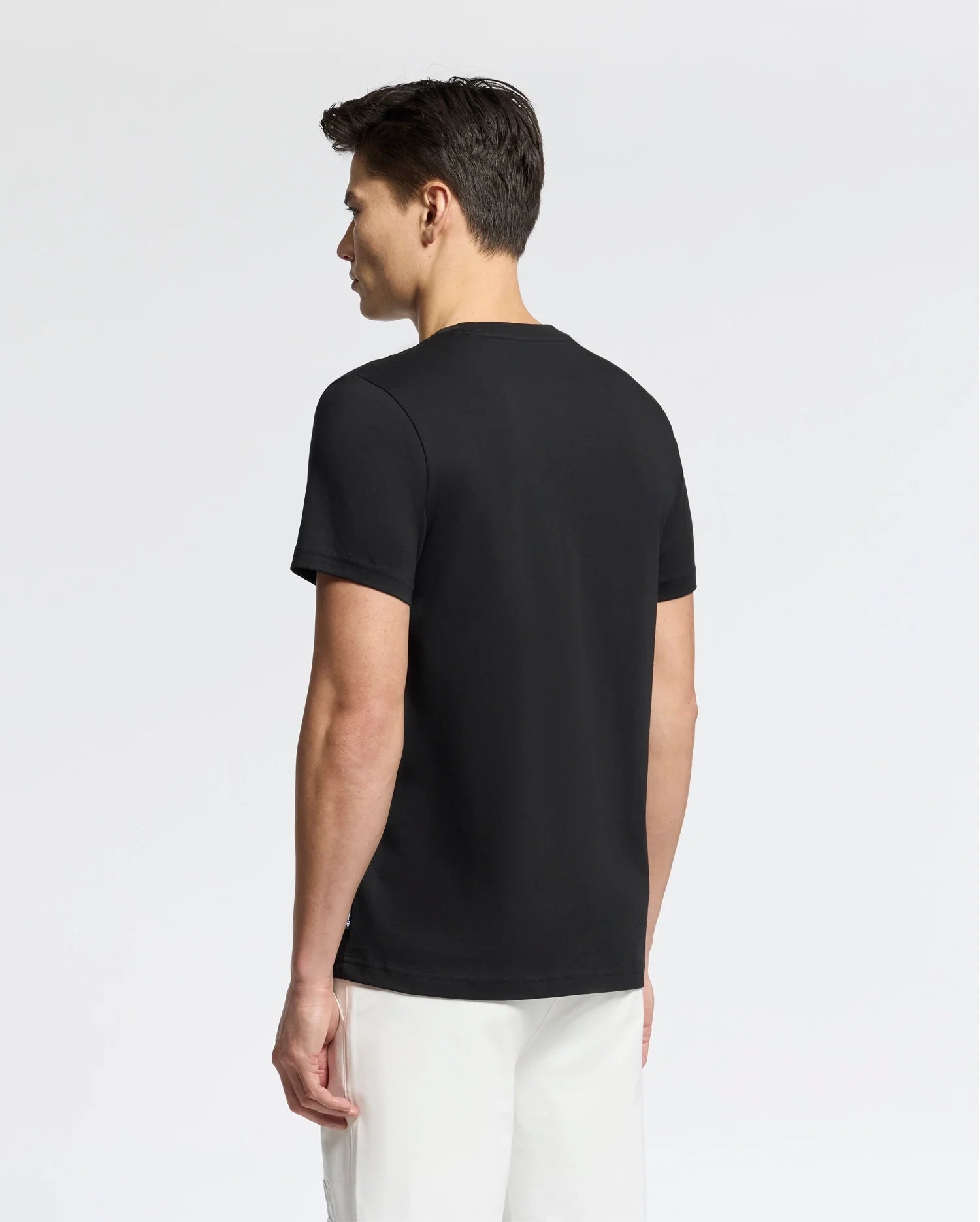 A person with short dark hair stands with their back to the camera, wearing a Psycho Bunny MENS SYDNEY WORDMARK GRAPHIC TEE in black and white pants against a plain white background.