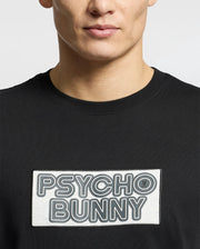 The individual wears the Psycho Bunny MENS SYDNEY WORDMARK GRAPHIC TEE, B6U510F200, a sleek black Pima cotton T-shirt with a rectangular "Psycho Bunny" patch in bold water-based print against a minimalist white background.