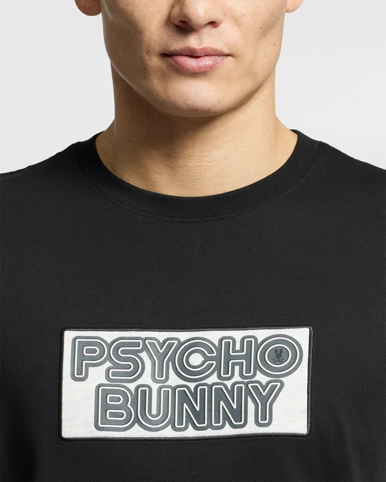 The individual wears the Psycho Bunny MENS SYDNEY WORDMARK GRAPHIC TEE, B6U510F200, a sleek black Pima cotton T-shirt with a rectangular "Psycho Bunny" patch in bold water-based print against a minimalist white background.