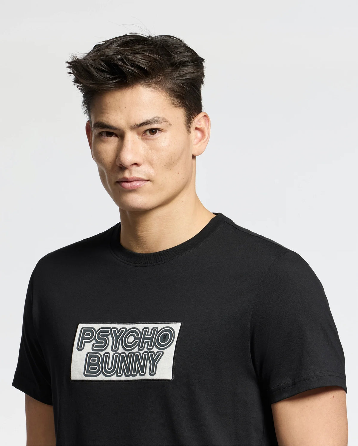 A person with short dark hair wears the MENS SYDNEY WORDMARK GRAPHIC TEE - B6U510F200 by Psycho Bunny, featuring the brand's text in a water-based print on the front. The background is plain light gray.