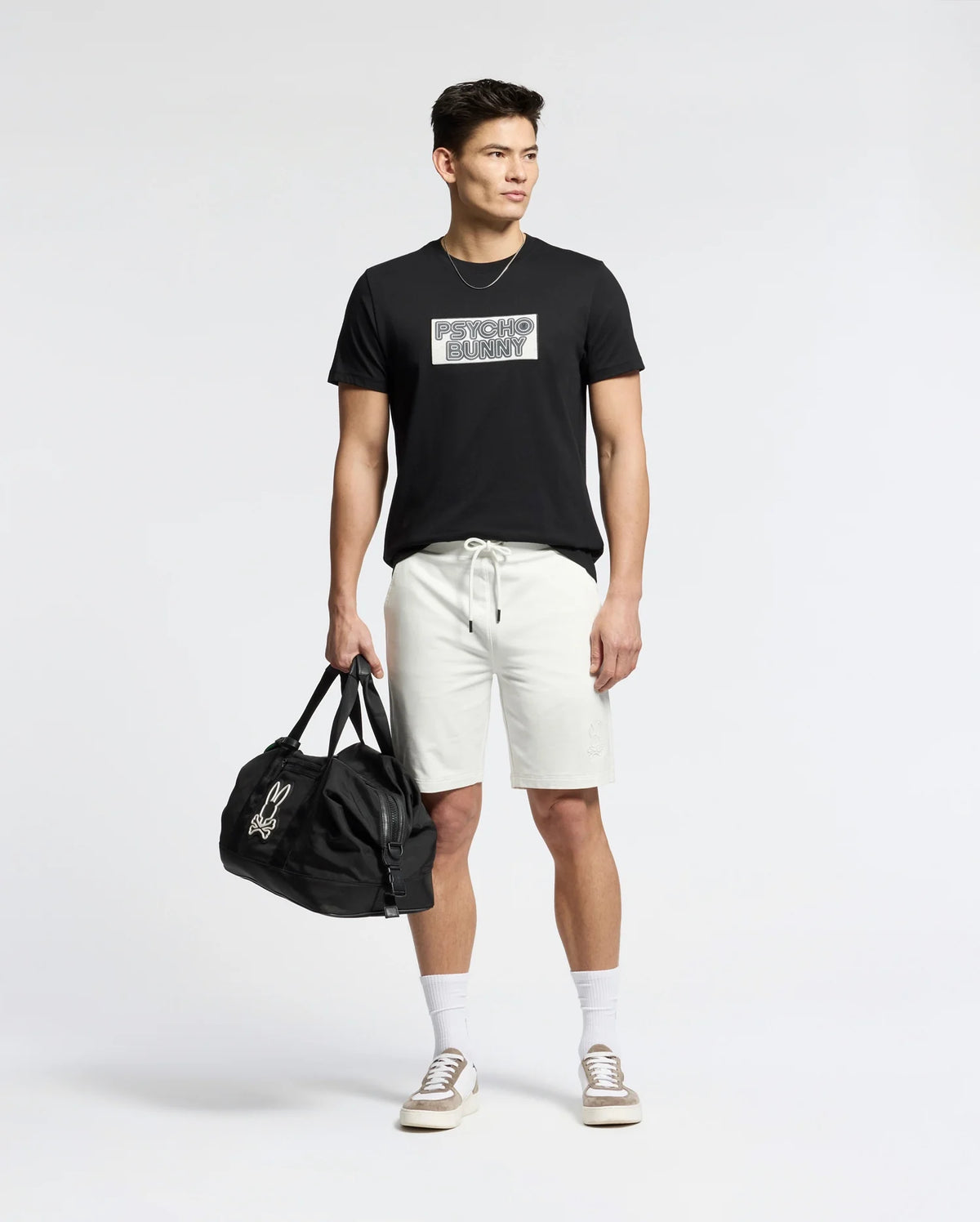 A person stands wearing a Psycho Bunny black Pima cotton MENS SYDNEY WORDMARK GRAPHIC TEE, white shorts, and beige sneakers. They hold a black duffel bag in their left hand against a plain white background.