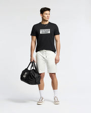 A person stands wearing a Psycho Bunny black Pima cotton MENS SYDNEY WORDMARK GRAPHIC TEE, white shorts, and beige sneakers. They hold a black duffel bag in their left hand against a plain white background.