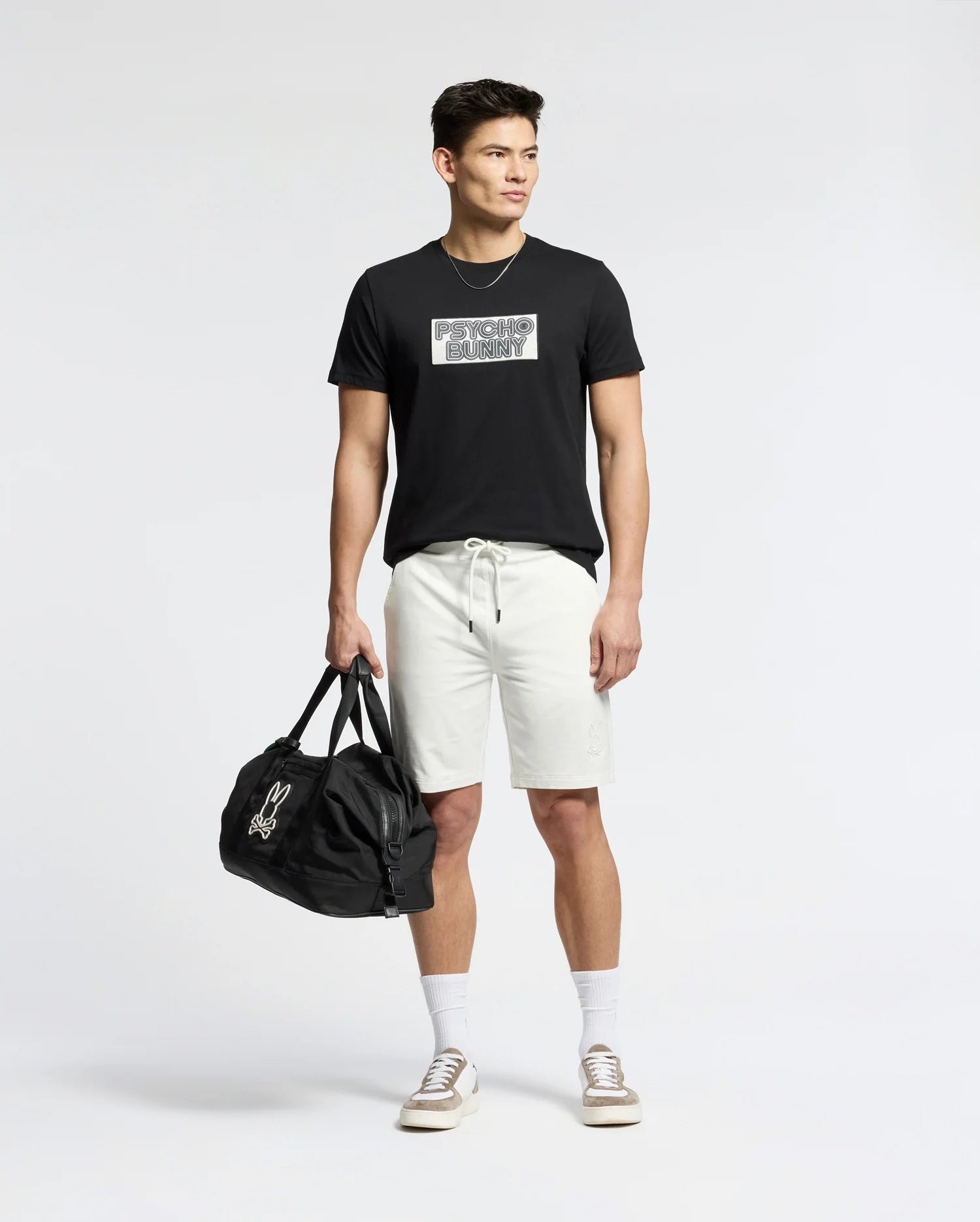 A person stands wearing a Psycho Bunny black Pima cotton MENS SYDNEY WORDMARK GRAPHIC TEE, white shorts, and beige sneakers. They hold a black duffel bag in their left hand against a plain white background.