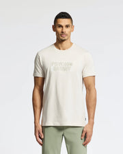 A man in a light-colored Psycho Bunny MENS SYDNEY WORDMARK GRAPHIC TEE (B6U510F200) stands against a plain background. His short, dark hair complements his outfit, which includes light green pants.