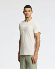 A man with short dark hair, against a plain backdrop, sports the MENS SYDNEY WORDMARK GRAPHIC TEE - B6U510F200 by Psycho Bunny, a light beige Pima cotton T-shirt. Paired with light green pants, he achieves a relaxed yet stylish look.
