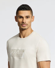 A man with short hair and a trimmed beard, looking serious, wears a luxurious light-colored MENS SYDNEY WORDMARK GRAPHIC TEE - B6U510F200 by Psycho Bunny. The plain, light background enhances his poised demeanor.