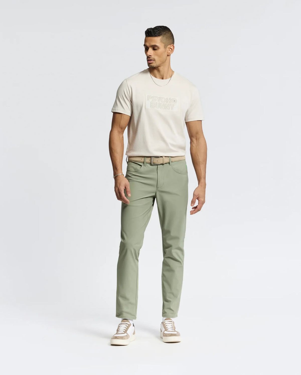 A person stands against a plain background in a light beige Psycho Bunny Mens Sydney Wordmark Graphic Tee (B6U510F200), paired with sage green pants and white sneakers. The wordmark graphic adds style as they maintain a neutral expression, looking slightly to the side.