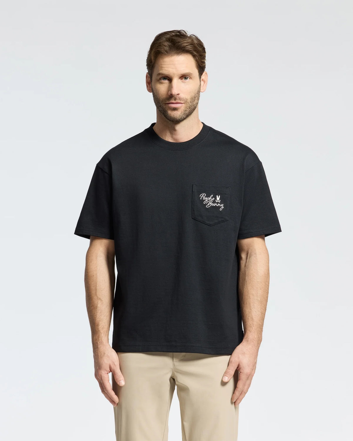 A man with short hair is wearing a Psycho Bunny MENS DESERT OVERSIZED POCKET TEE (B6U512F200) in black, featuring a white embroidery on the left chest pocket, paired with beige pants. The heavyweight Jersey tee's relaxed fit enhances his look as he stands against a plain, light gray background.