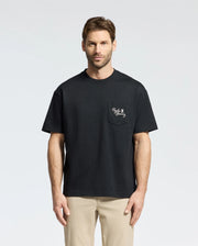 A man with short hair is wearing a Psycho Bunny MENS DESERT OVERSIZED POCKET TEE (B6U512F200) in black, featuring a white embroidery on the left chest pocket, paired with beige pants. The heavyweight Jersey tee's relaxed fit enhances his look as he stands against a plain, light gray background.