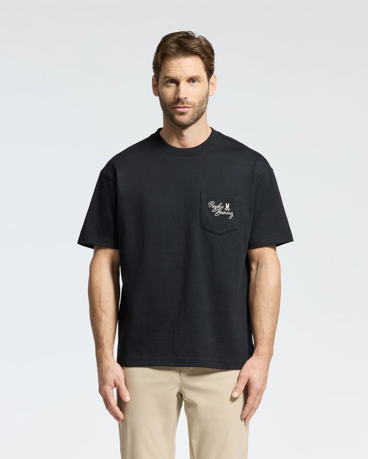 A man with short hair is wearing a Psycho Bunny MENS DESERT OVERSIZED POCKET TEE (B6U512F200) in black, featuring a white embroidery on the left chest pocket, paired with beige pants. The heavyweight Jersey tee's relaxed fit enhances his look as he stands against a plain, light gray background.