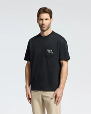 A man in a Psycho Bunny MENS DESERT OVERSIZED POCKET TEE - B6U512F200, crafted from Peruvian Pima cotton with a small graphic chest pocket, pairs it with beige pants against a light gray background.