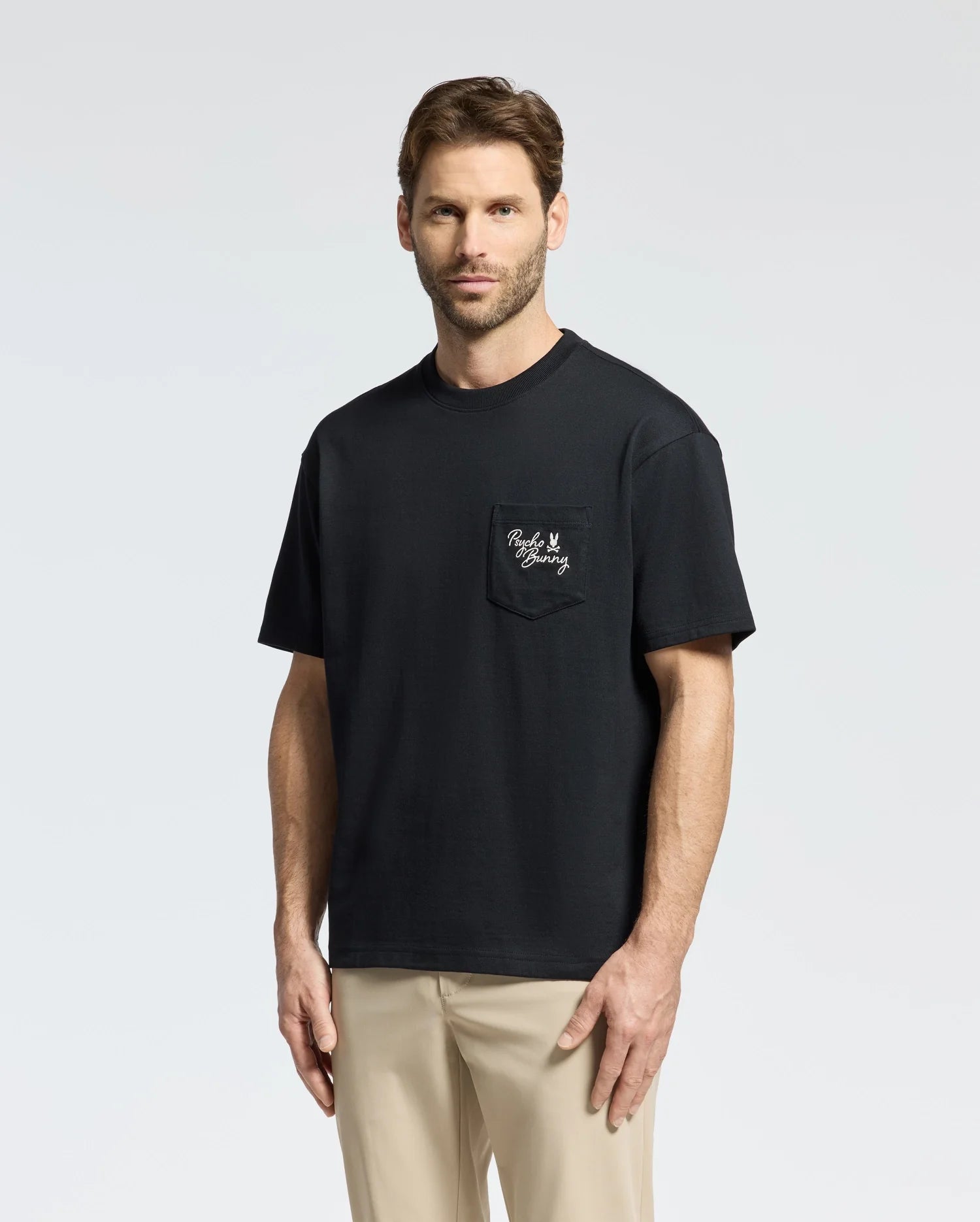 A man in a Psycho Bunny MENS DESERT OVERSIZED POCKET TEE - B6U512F200, crafted from Peruvian Pima cotton with a small graphic chest pocket, pairs it with beige pants against a light gray background.