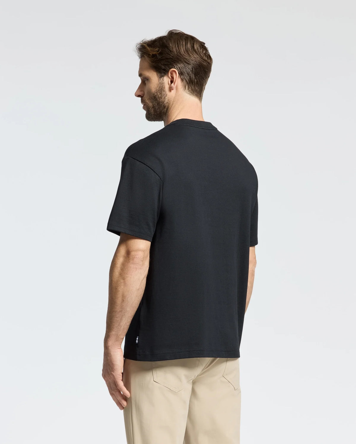 A man in a Psycho Bunny black MENS DESERT OVERSIZED POCKET TEE - B6U512F200, made from Peruvian Pima cotton, and beige pants stands sideways, gazing slightly downward. The relaxed fit enhances the casual vibe against a light-colored background.