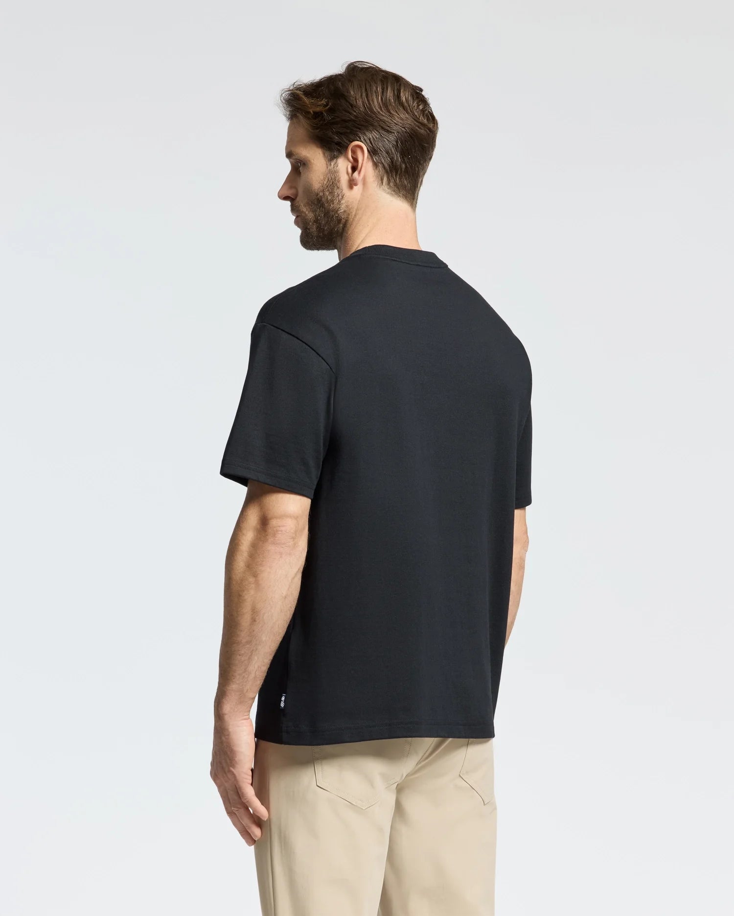 A man in a Psycho Bunny black MENS DESERT OVERSIZED POCKET TEE - B6U512F200, made from Peruvian Pima cotton, and beige pants stands sideways, gazing slightly downward. The relaxed fit enhances the casual vibe against a light-colored background.