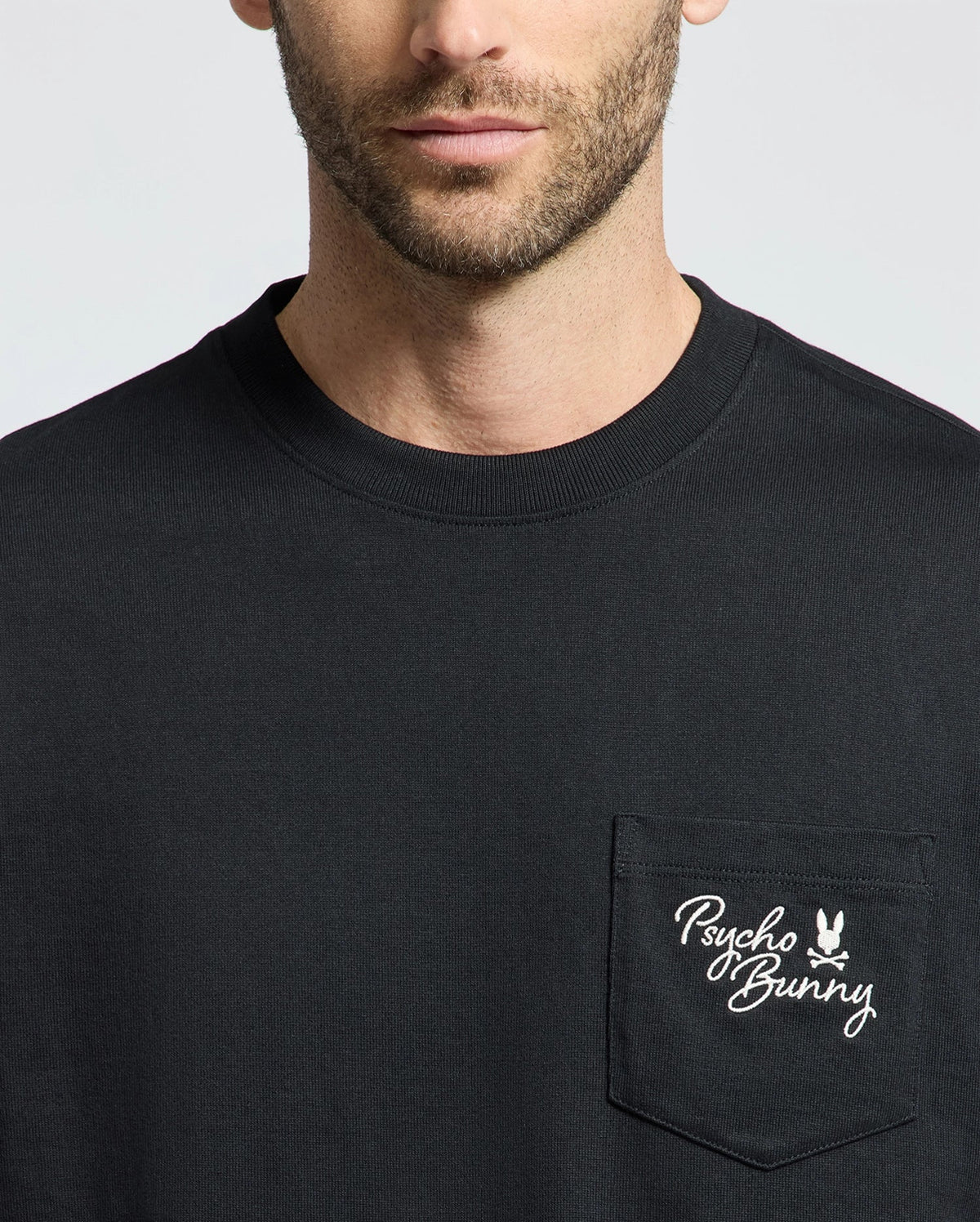 A person dons the Psycho Bunny MENS DESERT OVERSIZED POCKET TEE (B6U512F200) in black, showcasing a relaxed fit made of heavyweight Jersey. The chest pocket features a white Psycho Bunny logo with text and bunny graphic on a plain gray background.