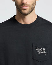 A person dons the Psycho Bunny MENS DESERT OVERSIZED POCKET TEE (B6U512F200) in black, showcasing a relaxed fit made of heavyweight Jersey. The chest pocket features a white Psycho Bunny logo with text and bunny graphic on a plain gray background.