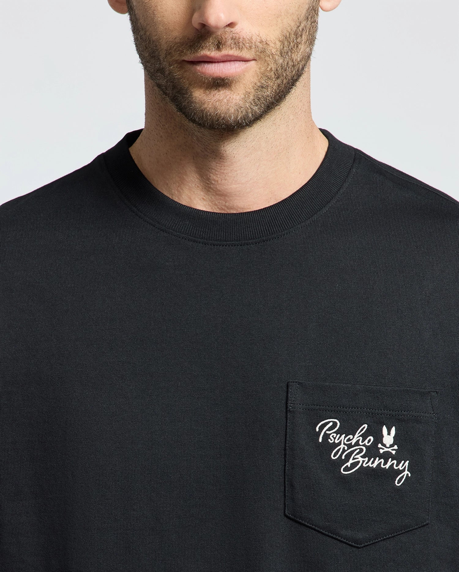 A person dons the Psycho Bunny MENS DESERT OVERSIZED POCKET TEE (B6U512F200) in black, showcasing a relaxed fit made of heavyweight Jersey. The chest pocket features a white Psycho Bunny logo with text and bunny graphic on a plain gray background.