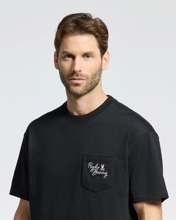 A man with short hair and a beard wears the Psycho Bunny MENS DESERT OVERSIZED POCKET TEE made from Peruvian Pima cotton. Featuring a relaxed fit, its black fabric contrasts nicely against the plain, light-colored background and includes a pocket with "Psycho Bunny" and an embroidered bunny logo in white.