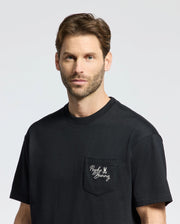 A man with short hair and a beard wears the Psycho Bunny MENS DESERT OVERSIZED POCKET TEE made from Peruvian Pima cotton. Featuring a relaxed fit, its black fabric contrasts nicely against the plain, light-colored background and includes a pocket with "Psycho Bunny" and an embroidered bunny logo in white.