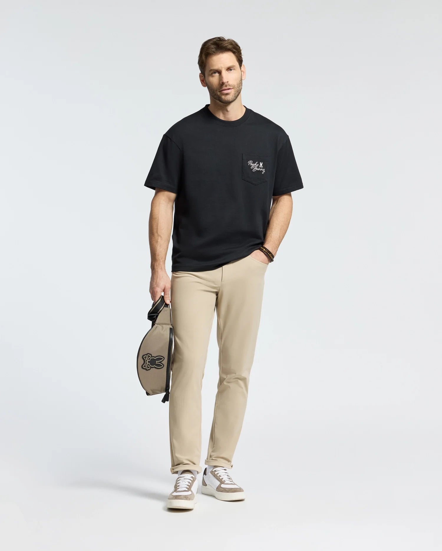 A man in a Psycho Bunny MENS DESERT OVERSIZED POCKET TEE (B6U512F200), beige pants, white sneakers, and a wristwatch stands holding a small bag against a plain, light backdrop that enhances his heavyweight jersey outfit perfectly.