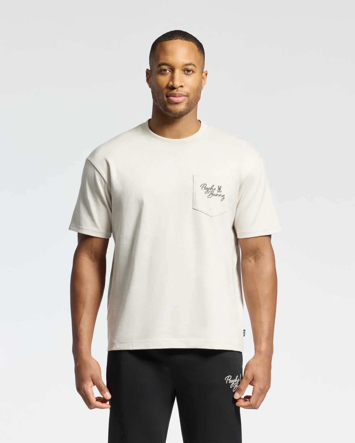 A man wears the Psycho Bunny MENS DESERT OVERSIZED POCKET TEE - B6U512F200, a beige heavyweight jersey T-shirt made from Pima cotton. It has a small text-designed pocket and is paired with black pants. He stands against a plain background with a neutral expression, highlighting its relaxed fit.