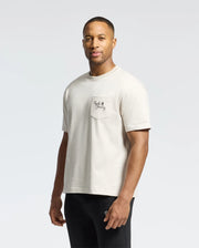 A man with short hair and a beard wears the Psycho Bunny MENS DESERT OVERSIZED POCKET TEE - B6U512F200 in relaxed-fit light beige Pima cotton, featuring a logo and text on a left-side pocket, paired with black pants against a plain white background.