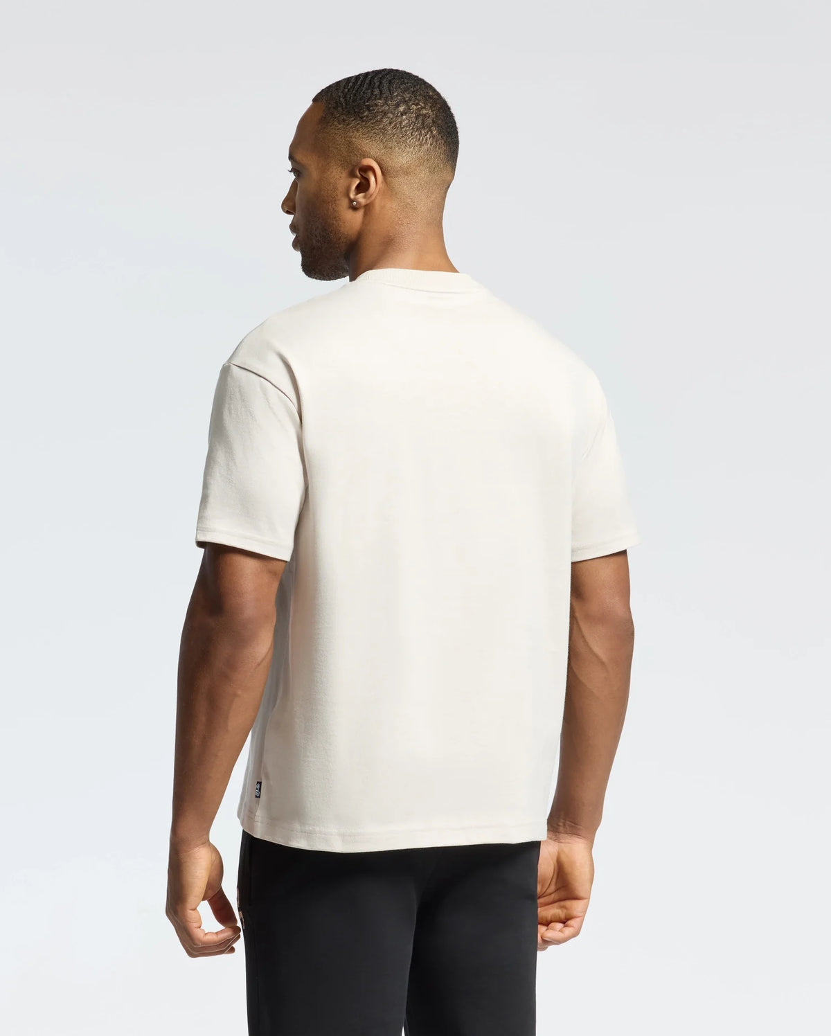 A person wearing a Psycho Bunny MENS DESERT OVERSIZED POCKET TEE - B6U512F200 in plain beige and black pants stands facing away, glancing to the side against a simple, light grey gradient background.
