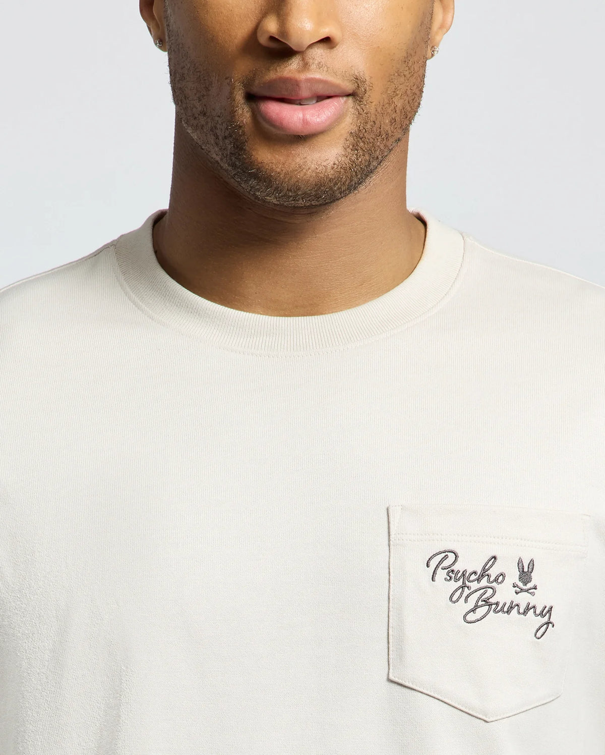 A man wears the Psycho Bunny Men's Desert Oversized Pocket Tee (B6U512F200) in light beige, featuring a chest pocket with the logo in dark lettering. The heavyweight jersey fabric provides a relaxed fit, exuding understated elegance with only the lower half of his face visible.