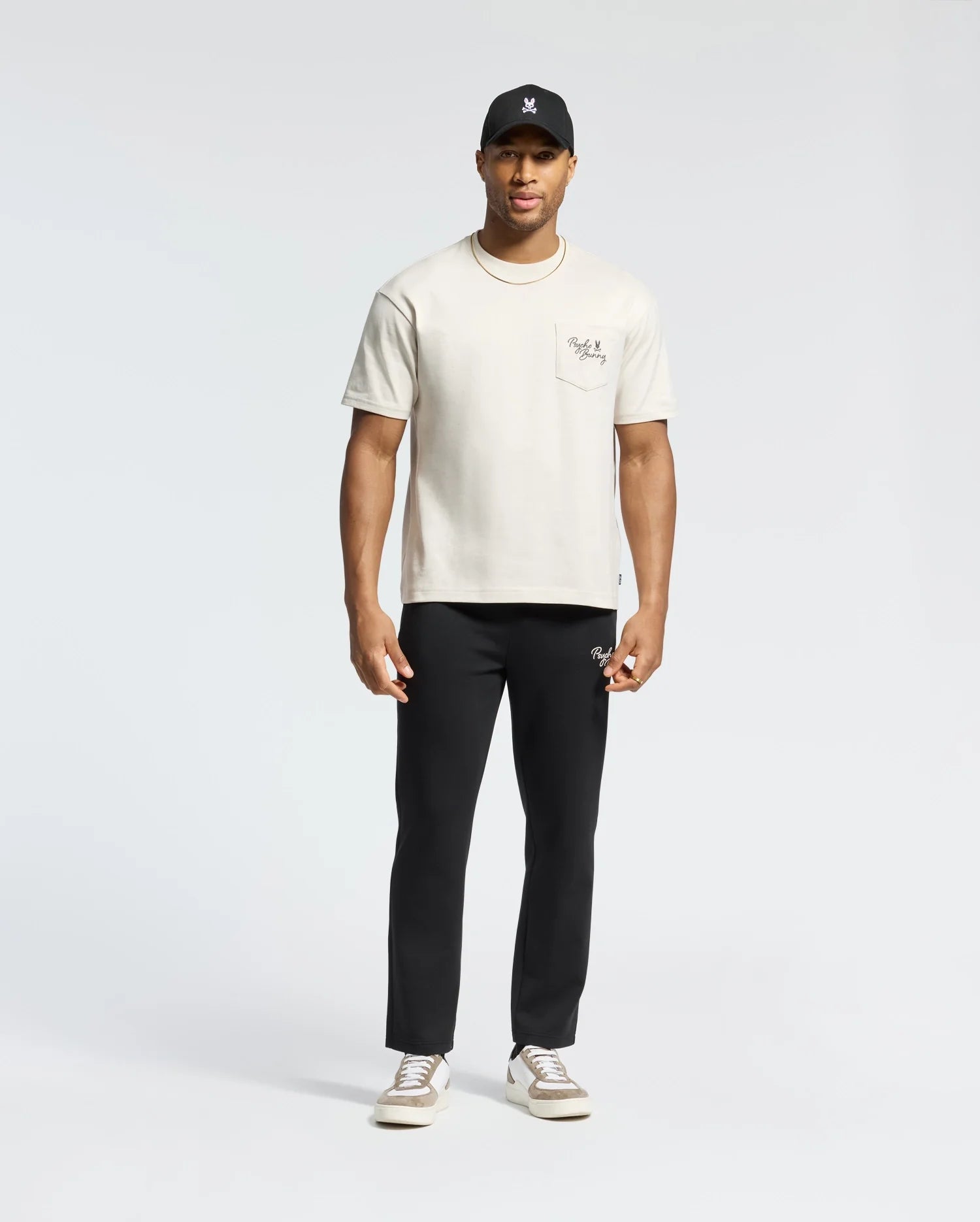 A man stands against a plain background wearing the MENS DESERT OVERSIZED POCKET TEE - B6U512F200 by Psycho Bunny, made from heavyweight Pima cotton, in beige, paired with black pants, a black cap, and white sneakers.