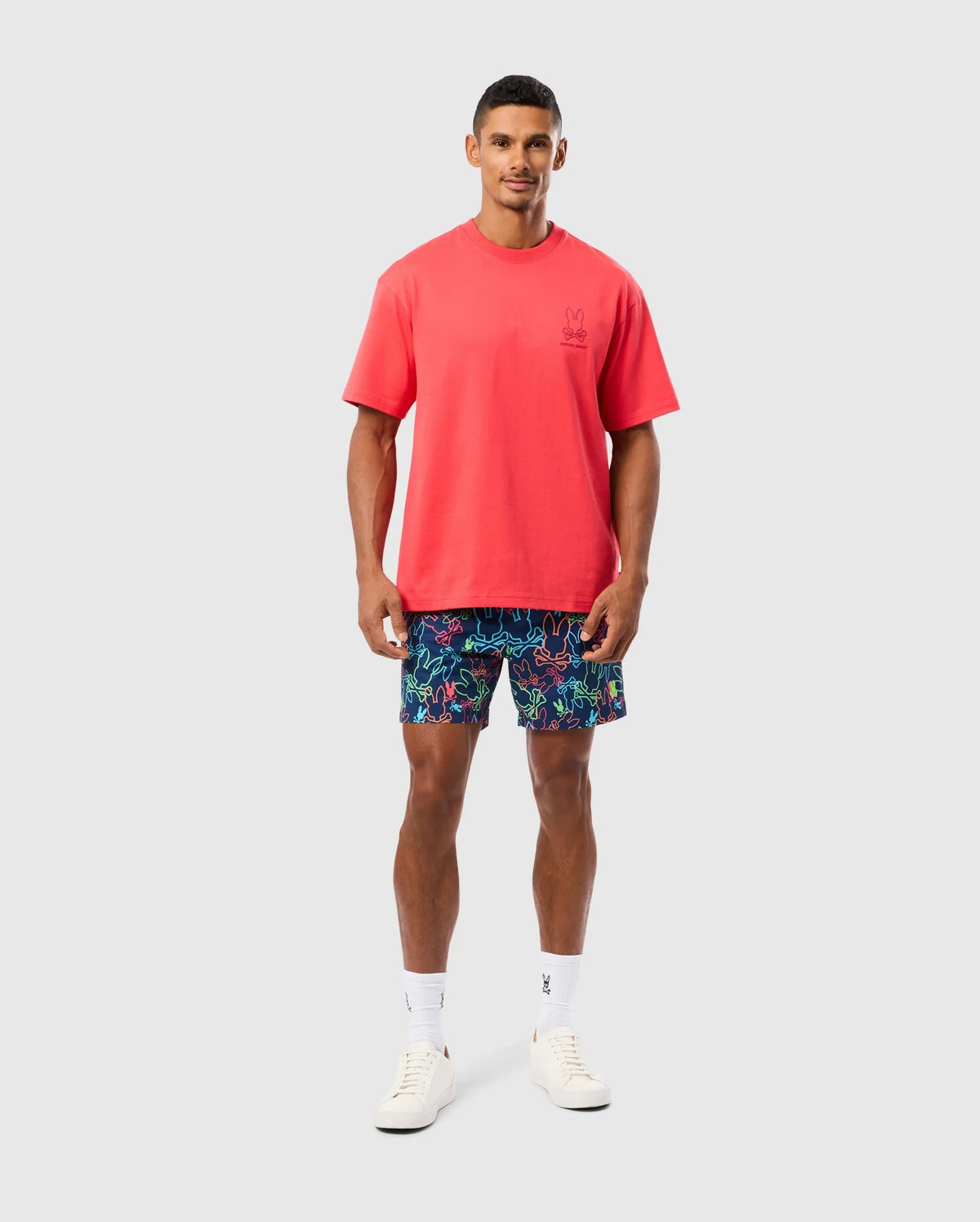 A person with short hair is wearing a bright red Psycho Bunny MENS BARRETT RELAXED FIT TEE - B6U518C200 featuring a Bunny graphic, multicolored shorts with a vibrant pattern, white socks, and white sneakers. The relaxed fit ensures comfort against the plain, light-colored background.