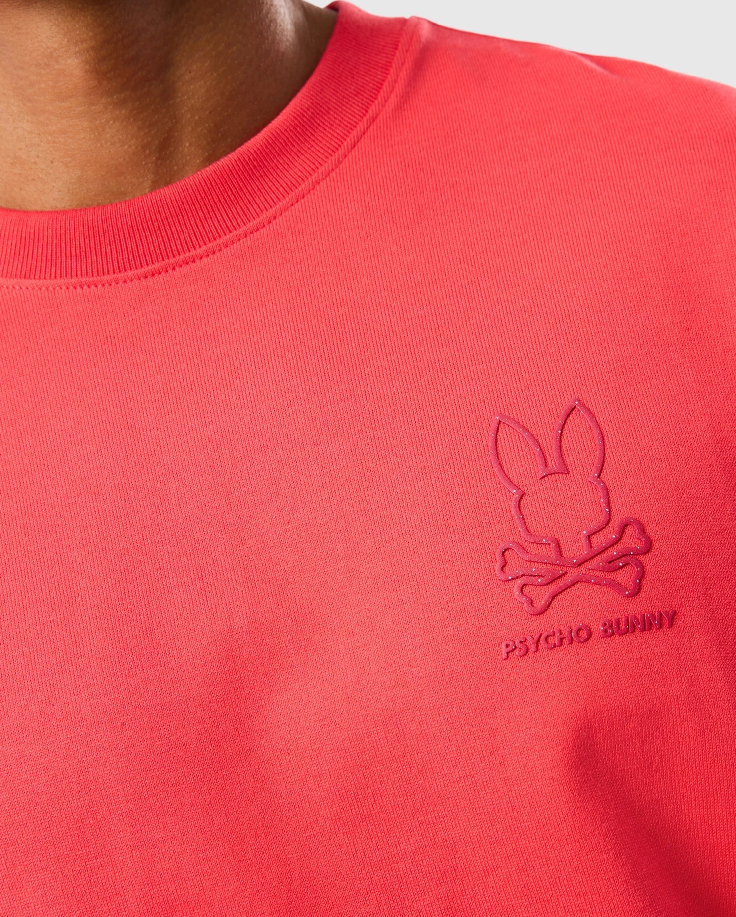 Close-up of a person wearing a relaxed fit red shirt with the Psycho Bunny logo embroidered on the chest. The logo features a minimalist rabbit head above a pair of crossed bones, showcasing impeccable style in heavyweight Pima cotton. This is the MENS BARRETT RELAXED FIT TEE - B6U518C200 by Psycho Bunny.