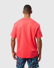 A person with short hair stands facing away, wearing a bright red Psycho Bunny MENS BARRETT RELAXED FIT TEE - B6U518C200 adorned with a subtle bunny graphic and colorful shorts featuring a pattern of various shapes and colors on a light grey background.