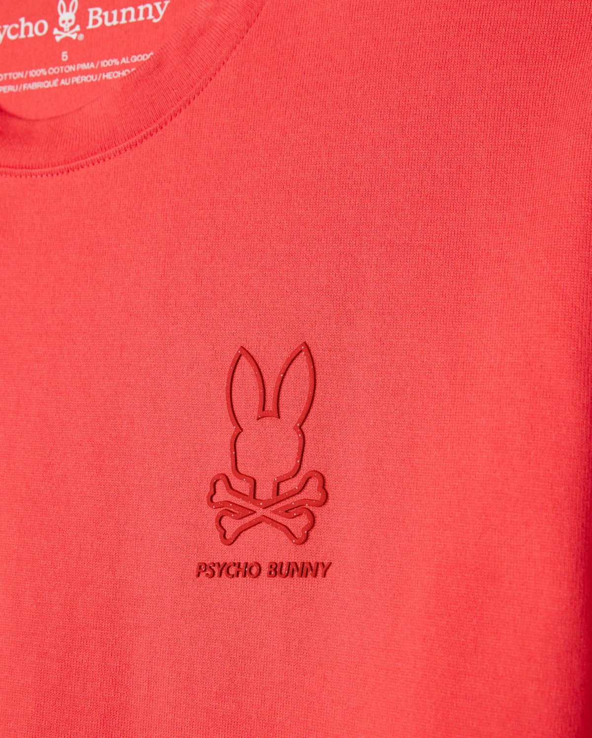 Close-up of a red Psycho Bunny MENS BARRETT RELAXED FIT TEE - B6U518C200 featuring an embossed Bunny graphic with a bunny head and crossbones below it. The text "PSYCHO BUNNY" is stitched under the logo. Made from smooth, heavyweight Pima cotton, the fabric ensures durability while maintaining a relaxed fit.
